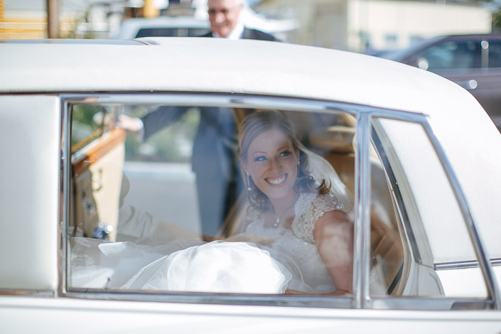 The bride's smile fills the window of the cream vintage car, a man in grey outside.
