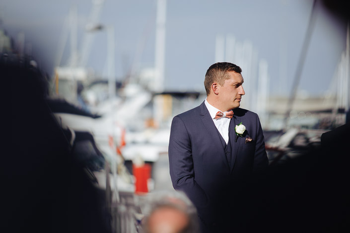 The groom waits between the blurred shoulders, masts crowding behind
