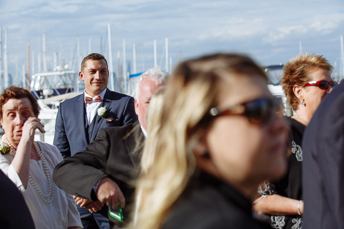 A white haired woman in pearls dabs her eye as the groom watches the aisle.