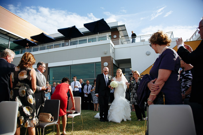 Down the clubhouse lawn the bride comes on the arm of the grey haired man in glasses.