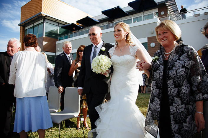 A woman in a leaf print jacket beams as the bride's walk reaches the water's edge.