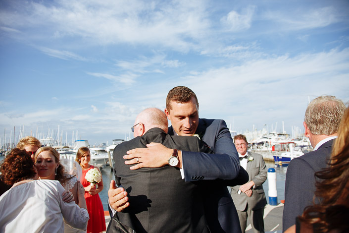 A crushing hug between the groom and a balding man in grey on the boardwalk.