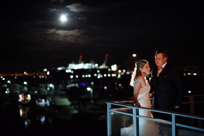 Moonlight over the marina as the bride and groom lean at the balcony rail.