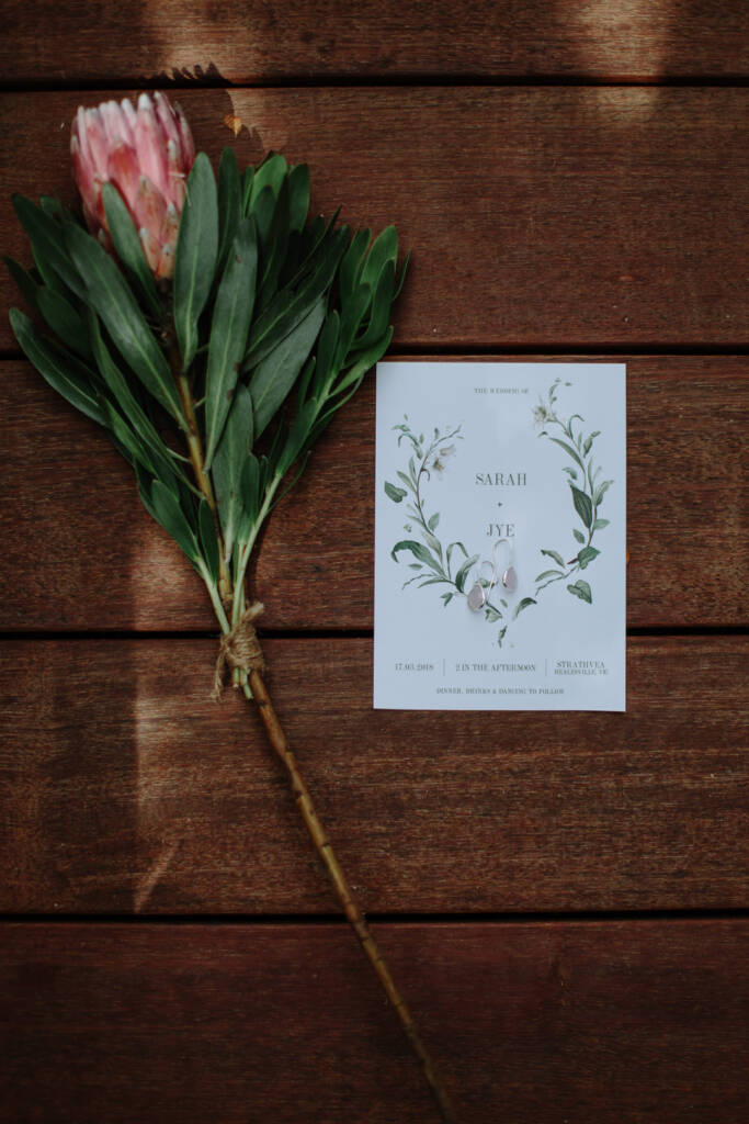 Sarah and Jye's Strathvea invitation lies beside earrings and a pink protea on a timber surface.
