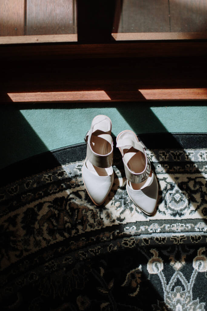 Pale pointed bridal shoes with metallic straps rest on a patterned rug.