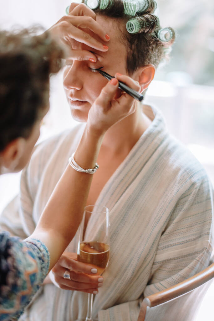 Sarah has her eye make-up applied while wearing hair rollers and holding a flute.