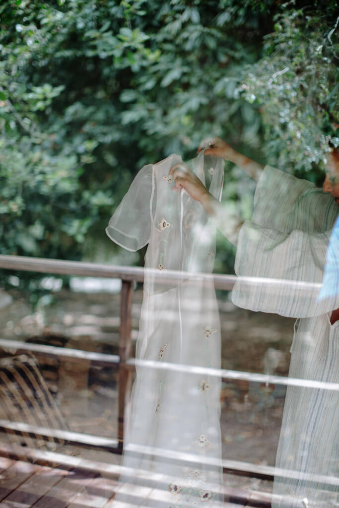 Sarah holds her embroidered gown behind glass reflecting the trees at Strathvea Guesthouse.