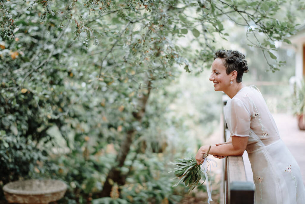 Sarah leans on the timber verandah railing at Strathvea, holding her bouquet towards the garden.