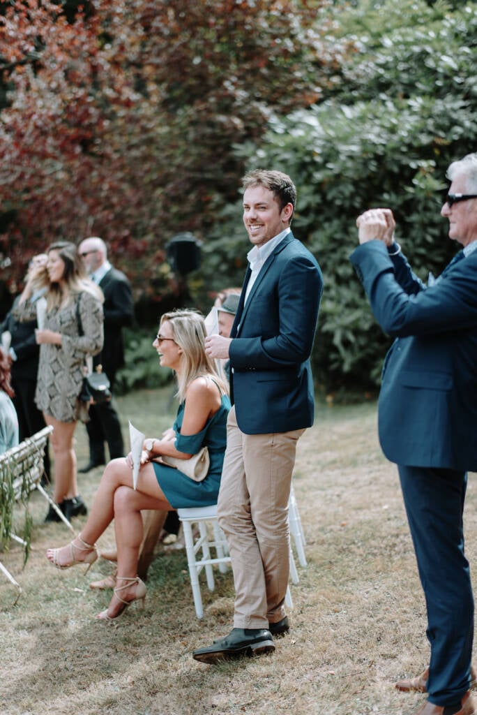 A guest smiles while holding a sheet of paper as another photographs the garden ceremony at Strathvea.