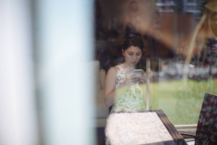Through glass, Shannon in a floral dress looks down at her phone, the room reflected across the pane.