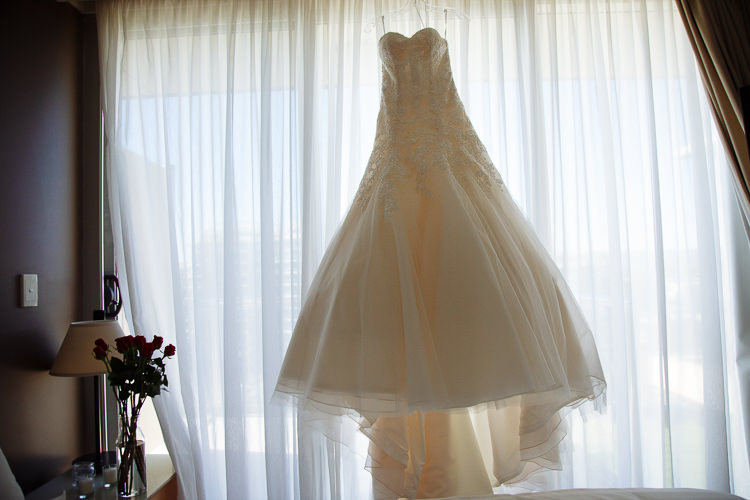 A strapless lace wedding gown hangs before sheer curtains, with red roses on the bedside table.