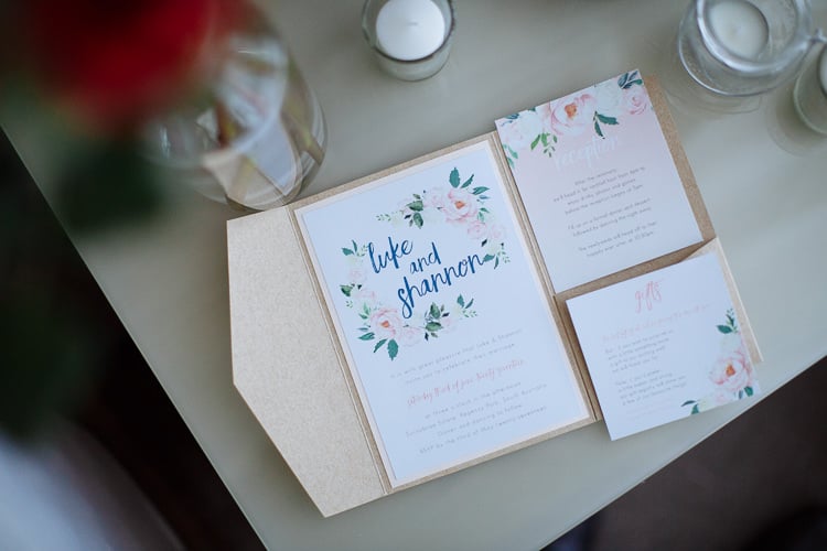 A floral invitation suite lettered Luke and Shannon for Sunnybrae Estate in a gold envelope beside candles.
