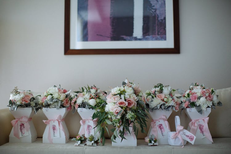Six pink and white rose bouquets in labelled white boxes line a sofa with a flower girl basket.