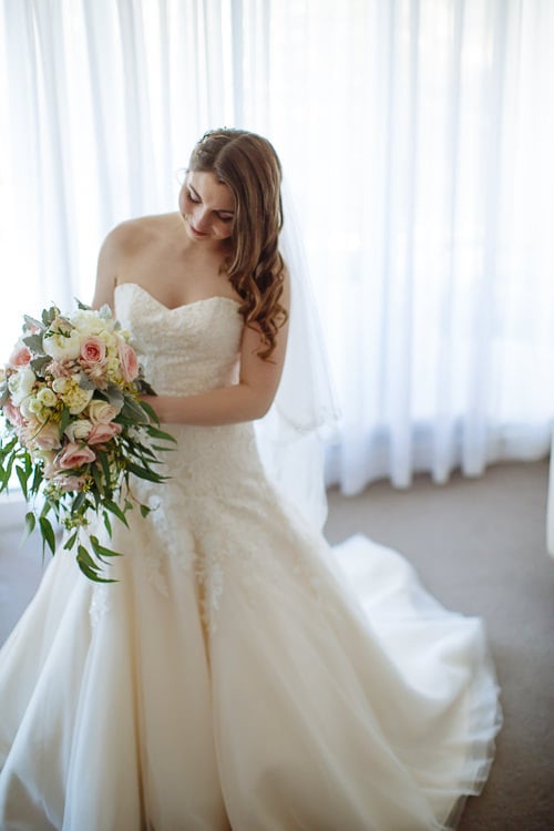 Full-length portrait of Shannon in her strapless lace wedding gown before sheer curtains, holding her bouquet.
