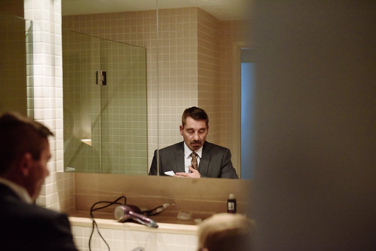 In a tiled bathroom mirror, a man with a moustache in a grey suit checks his pocket square.