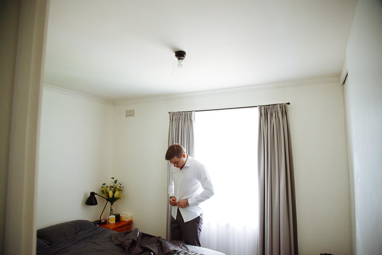 Luke buttons his white shirt beside a bedroom window.