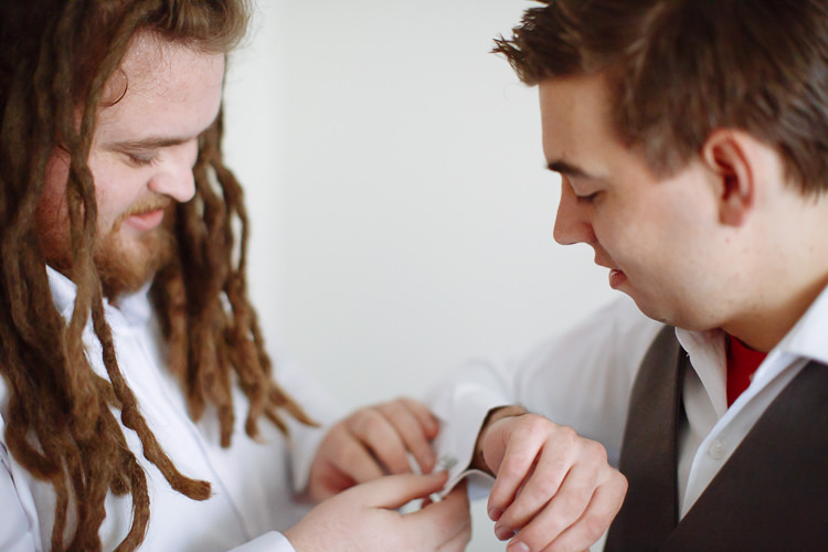 A groomsman with long dreadlocks fastens Luke's cufflink as Luke looks down in his charcoal waistcoat.