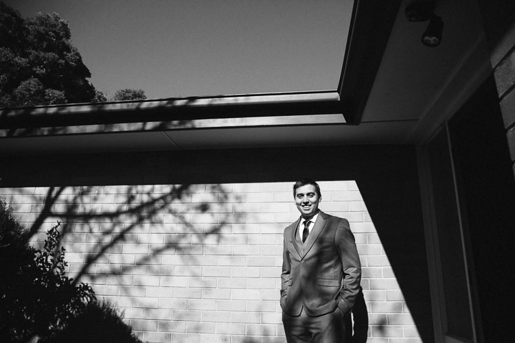 Black and white of Luke standing hands in pockets against a sunlit brick wall beneath a deep eave.
