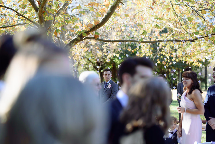 The groom waits beneath leafy branches at Sunnybrae, seen between guests in the foreground.