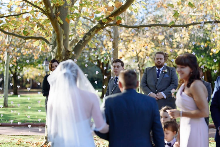 Luke watches Shannon approach beneath paper hearts and leafy branches at Sunnybrae.