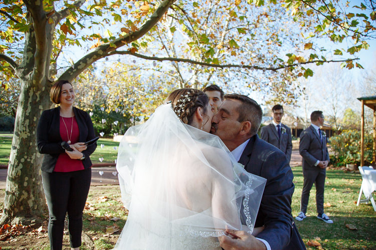 The man with the moustache kisses Shannon's cheek beneath the lifted veil, the celebrant smiling.