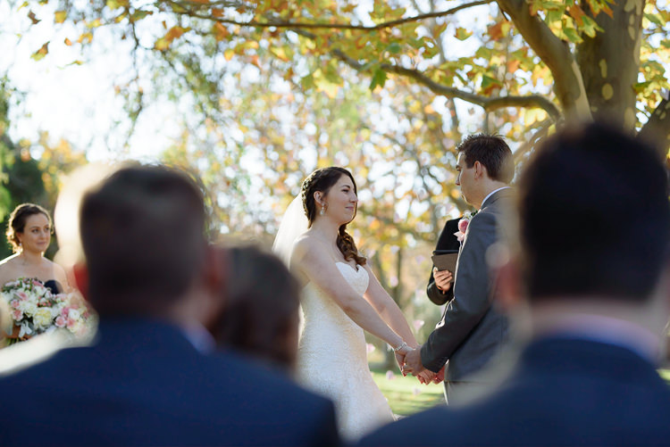 Shannon and Luke hold hands beneath yellow-green leaves during their garden wedding ceremony at Sunnybrae Estate.