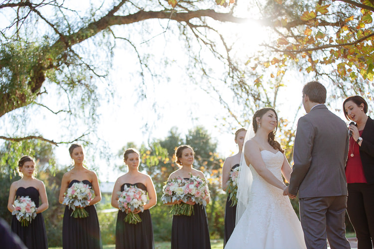 Shannon and Luke hold hands as the celebrant speaks beneath sunlit branches at Sunnybrae.