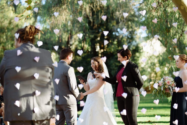 Shannon reads from a tablet as the celebrant holds a microphone at Sunnybrae Estate, paper hearts hanging nearby.