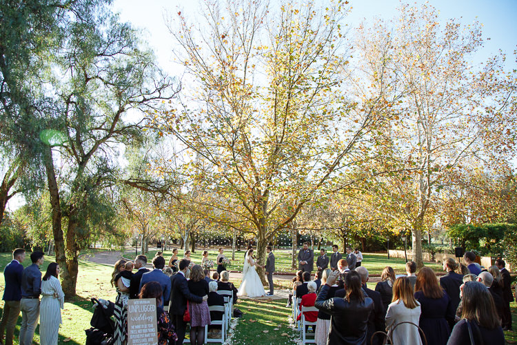 Guests watch the garden wedding ceremony beneath trees at Sunnybrae, with an unplugged wedding sign beside the aisle.