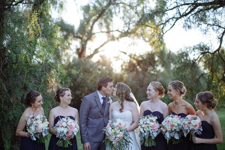 Shannon and Luke kiss between five bridesmaids beneath sunlit trees at Sunnybrae.