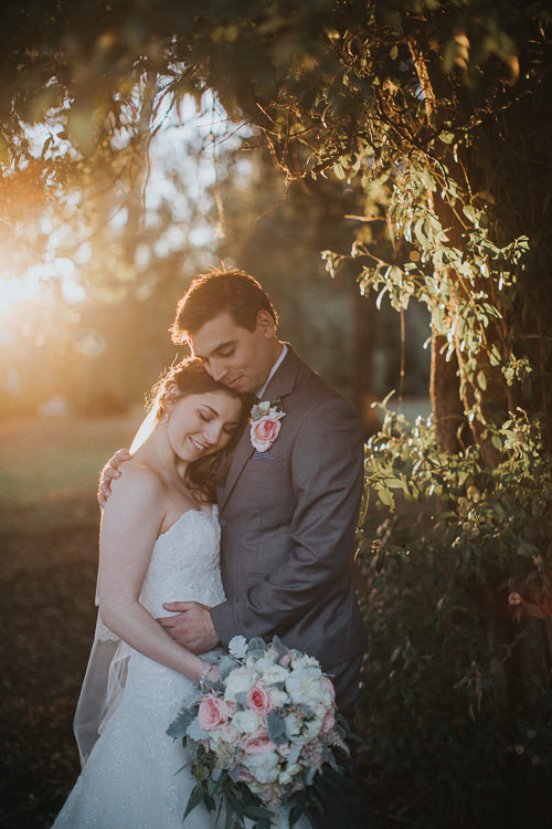 The couple embrace beneath leafy branches at Sunnybrae Estate, with warm light behind them.