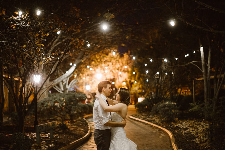 Shannon and Luke embrace at night on a garden path at Sunnybrae Estate beneath festoon lights.