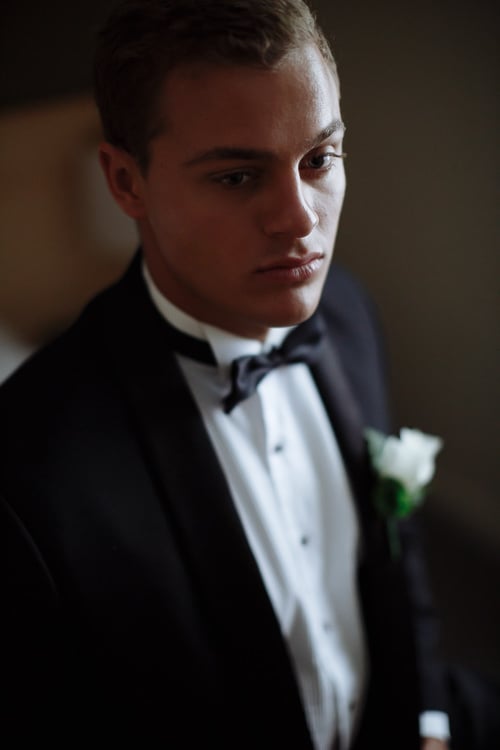 Close portrait of the man in a shawl collar tuxedo looking down, white rose on the lapel.
