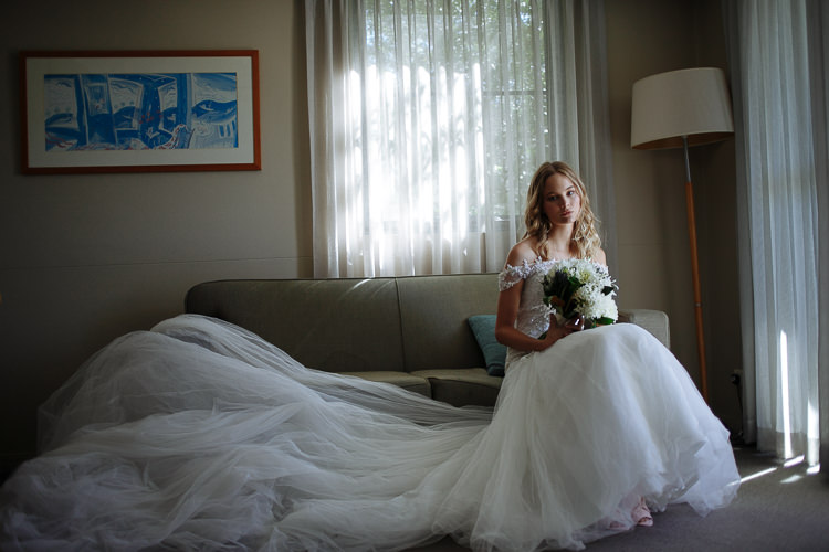 The woman in the off shoulder lace gown sits by sheer curtains, the tulle train drowning the couch.