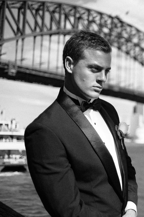 Black-and-white portrait of a man on the Sydney Harbour waterfront beneath the Bridge, with a ferry behind.