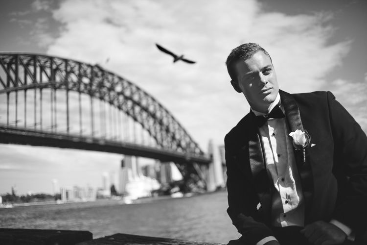 A gull flies above a man in a tuxedo beside Sydney Harbour, with the Bridge arch behind.