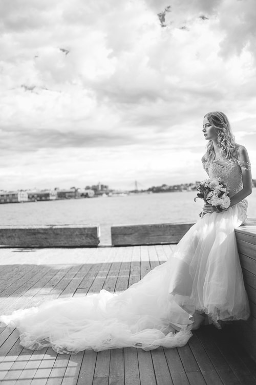 A woman sits above Sydney Harbour, her gown's train spread across the timber platform.