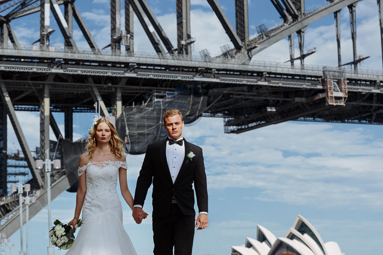 The couple walk hand in hand beside Sydney Harbour beneath the Bridge, with Opera House sails behind.