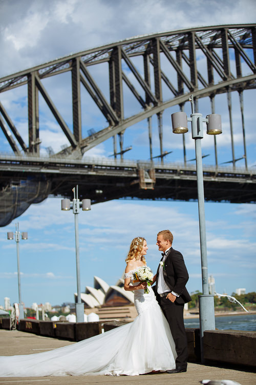 The couple laugh on a Sydney Harbour boardwalk beneath the Bridge, with Opera House sails beyond.