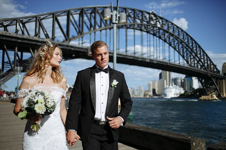 The couple walk beside Sydney Harbour beneath the Bridge, with a cruise ship across the water.
