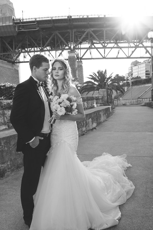 In black and white, the couple stand beside a stone wall on the Sydney Harbour waterfront beneath the Bridge.