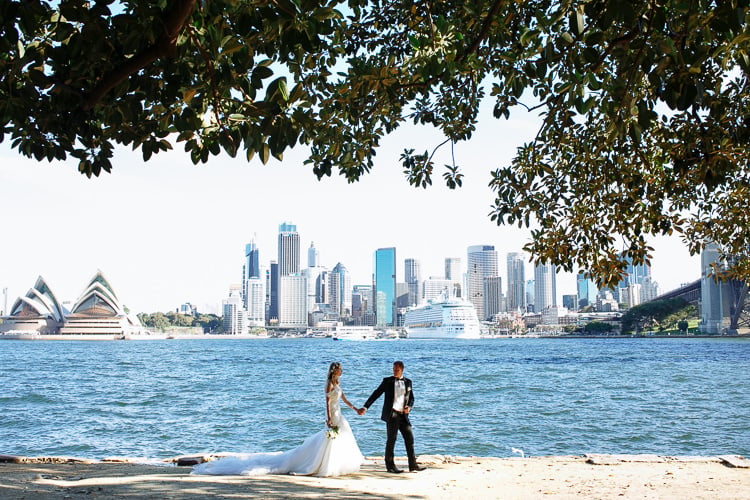 The couple hold hands on a sandy Sydney Harbour shore, with the Opera House and skyline across the water.