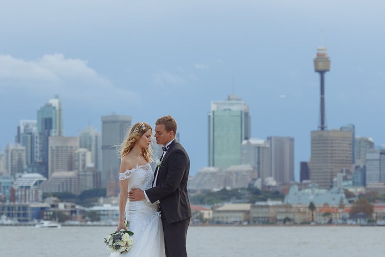The couple stand close beside Sydney Harbour, with Sydney Tower and the skyline across the water.