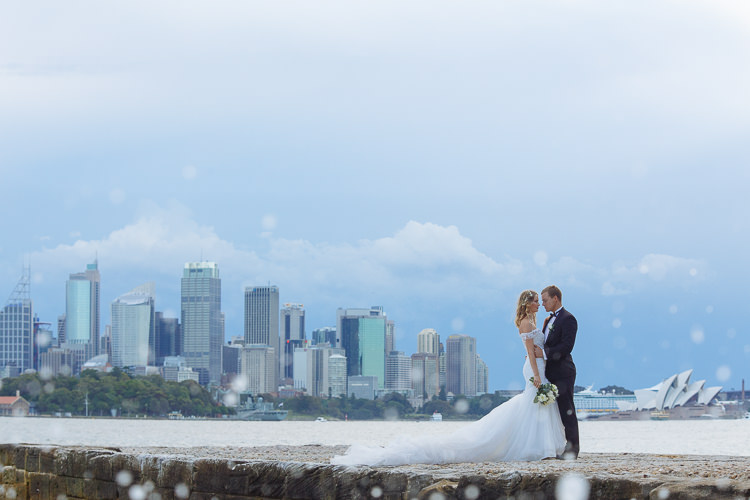 The couple stand on a stone seawall beside Sydney Harbour, with the Opera House and skyline across the water.