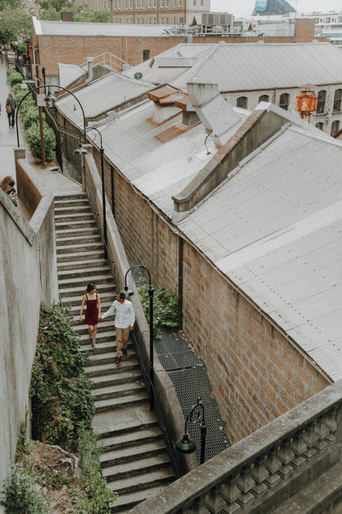 Jeannette and Pierre walk down sandstone steps in The Rocks, Sydney CBD, beside heritage buildings.