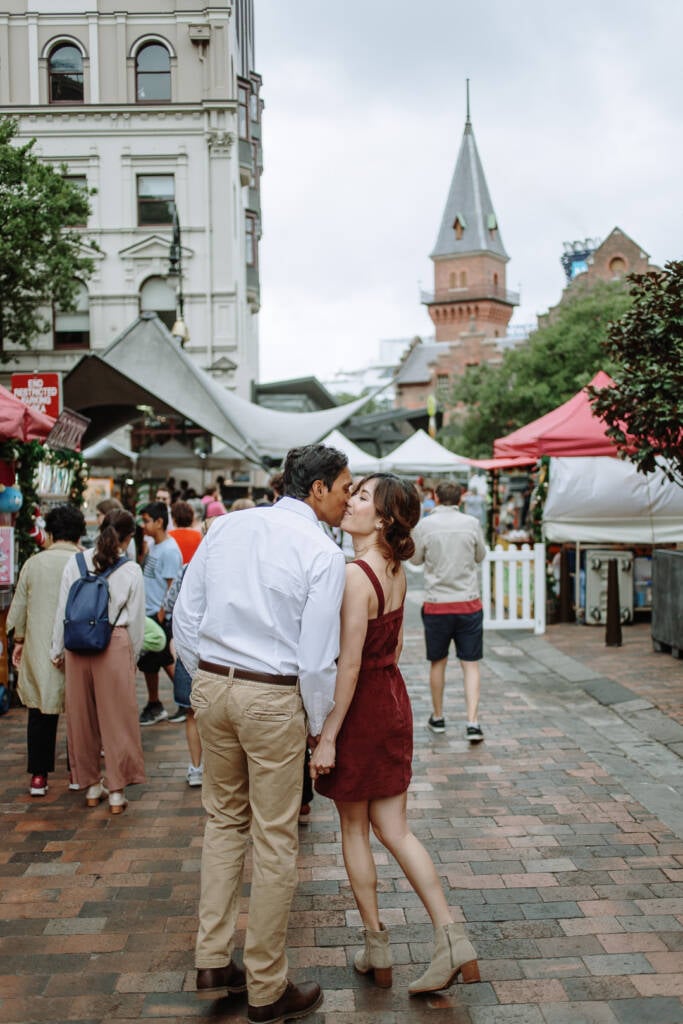 Jeannette and Pierre kiss beside the weekend market stalls at The Rocks Markets beneath a brick spire.