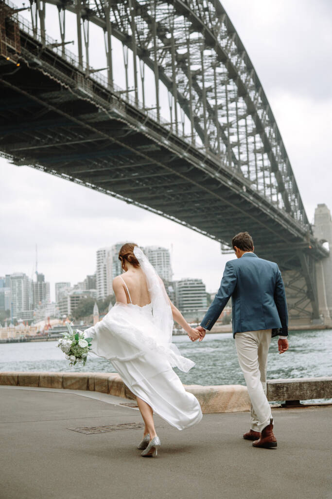 From behind, Jeannette and Pierre walk hand in hand near Pylon Lookout beneath Sydney Harbour Bridge.