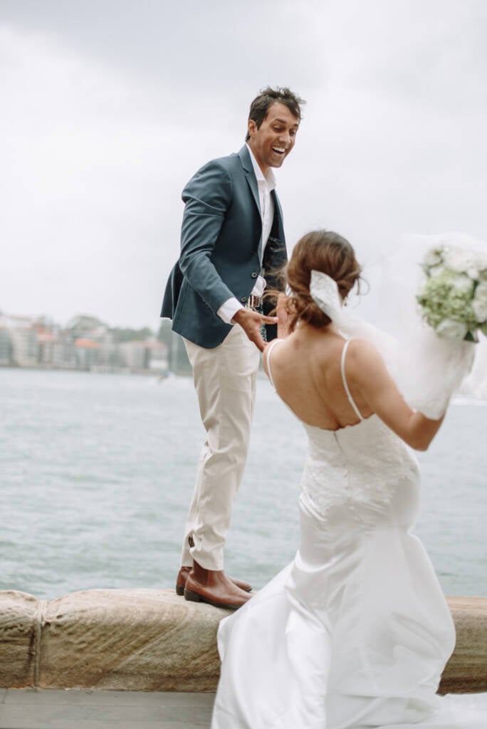 Near Hickson Road Reserve, Pierre laughs as he reaches down to Jeannette, who holds a white bouquet beside the sandstone harbour wall.