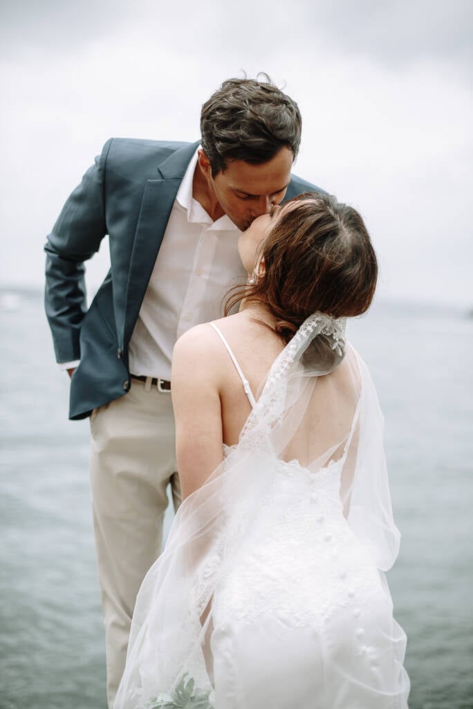 Pierre bends to kiss Jeannette near Hickson Road Reserve, her veil pinned beneath a low bun and the harbour behind.