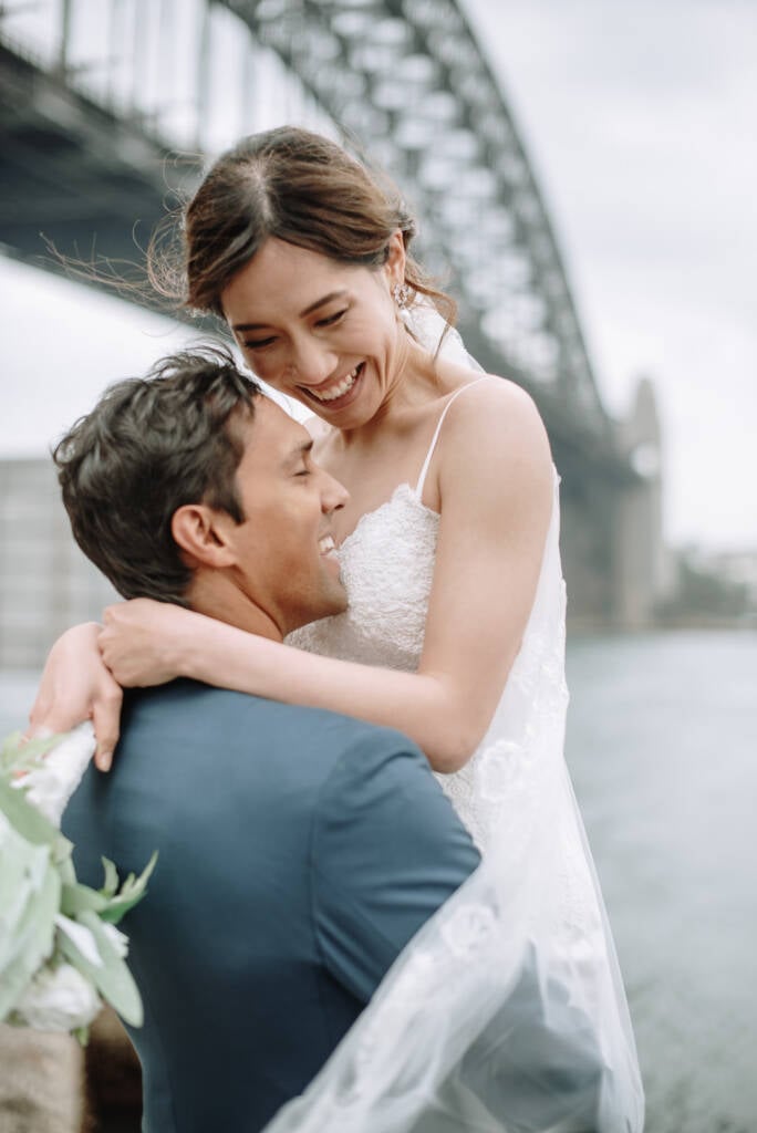 Jeannette laughs with her arms around Pierre near Hickson Road Reserve, Sydney Harbour Bridge behind them.